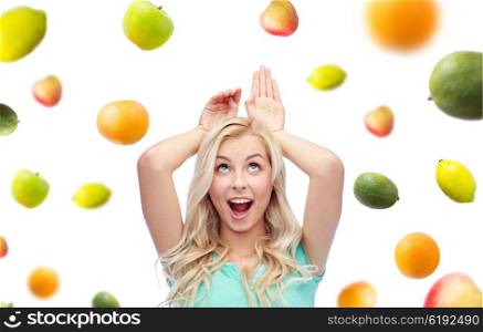 fun, expressions, healthy eating and people concept - happy smiling young woman making bunny ears over fruits on white background