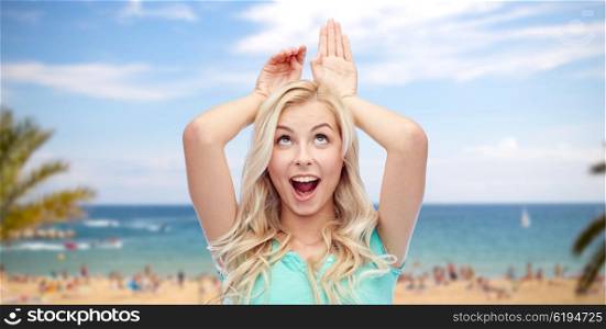 fun, expressions, summer holidays, travel and people concept - happy smiling young woman making bunny ears over exotic tropical beach with palm trees and sea background