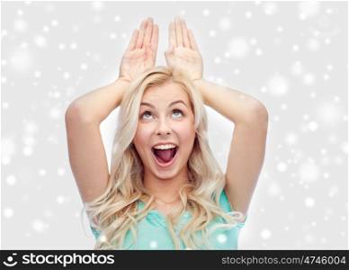 fun, expressions, winter holidays, christmas and people concept - happy smiling young woman making bunny ears over snow