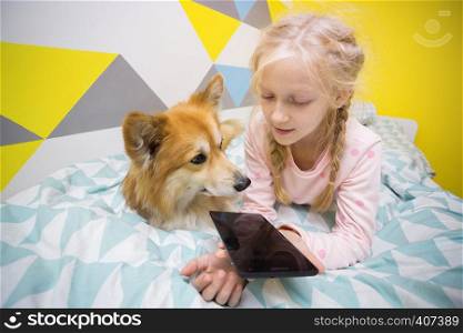 fun girl and dog corgi on the bed in the nursery play on the tablet