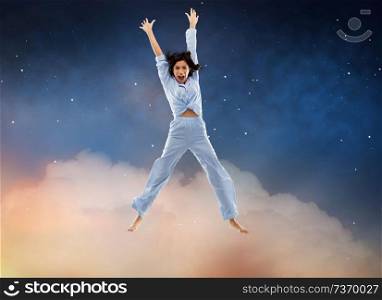 fun, people and bedtime concept - happy young woman full of energy in blue pajama jumping over starry night sky background. happy woman in blue pajama jumping high