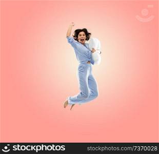 fun, people and bedtime concept - happy young woman full of energy in blue pajama holding pillow and jumping over living coral pink background. happy woman in blue pajama jumping with pillow