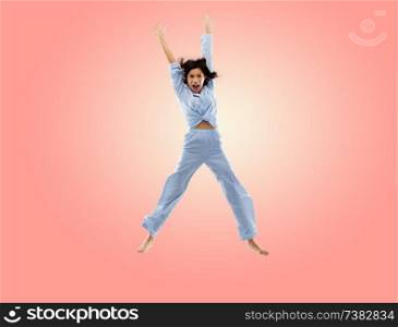 fun, people and bedtime concept - happy young woman full of energy in blue pajama jumping over living coral pink background. happy woman in blue pajama jumping high over pink