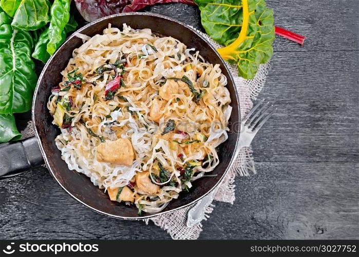 Funchoza with chard and meat in pan on board top. Rice noodles with leafy beet, chicken breast meat, cashew nuts and soy sauce in a frying pan on burlap on wooden plank background on top