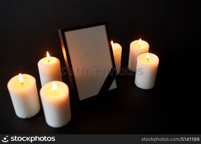 funeral and mourning concept - photo frame with black ribbon and burning candles over dark background. photo frame with black mourning ribbon and candles