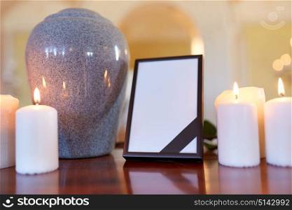 funeral and mourning concept - photo frame with black ribbon, cremation urn and burning candles on table in church. photo frame, cremation urn and candles in church
