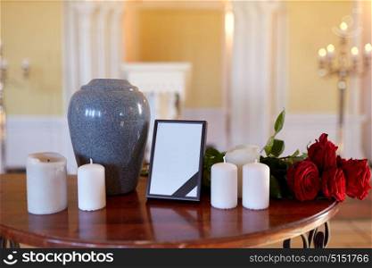 funeral and mourning concept - photo frame with black ribbon, cremation urn, flowers and candles on table in church. photo frame, cremation urn and candles in church