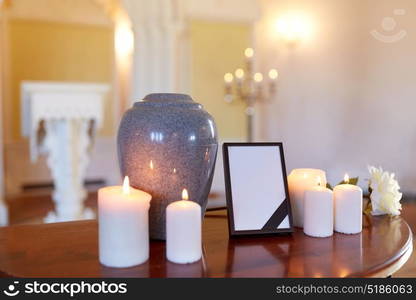 funeral and mourning concept - photo frame with black ribbon, cremation urn and burning candles on table in church. photo frame, cremation urn and candles in church