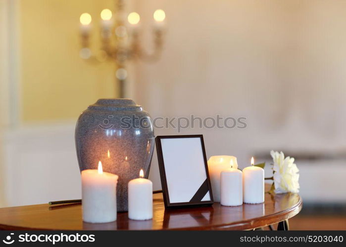 funeral and mourning concept - photo frame with black ribbon, cremation urn and burning candles on table in church. photo frame, cremation urn and candles in church