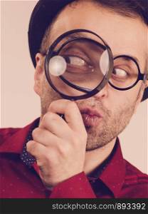 Funny adult guy wearig weird eyeglasses investigating something using magnifying glass making his eye looking big.. Funny man with magnifying glass