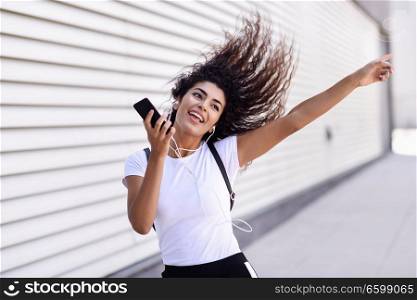 Funny African woman listening to music and dancing with earphones and smartphone outdoors. Arab girl in sport clothes with moving curly hairstyle in urban background.