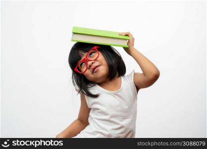 Funny and Happy Asian little preschool girl wearing red glasses holding a green book on the head, on white isolated background. Concept of school kid and education in elementary and preschool, home school