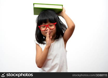 Funny and Happy Asian little preschool girl wearing red glasses holding a green book on the head, on white isolated background. Concept of school kid and education in elementary and preschool, home school