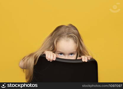 Funny and pretty child girl with long fair hair peeks out from behind a chair. the child is hiding behind a chair. Funny and pretty child girl with long fair hair peeks out from behind a chair.