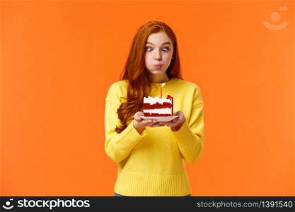 Funny and silly cute redhead b-day girl squinting eyes making goofy expression, pouting as holding birthday cake and blowing-out candle to make wish, having fun at party celebrating, orange wall.. Funny and silly cute redhead b-day girl squinting eyes making goofy expression, pouting as holding birthday cake and blowing-out candle to make wish, having fun at party celebrating, orange wall