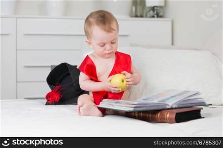 Funny baby in graduation cap and ribbon holding apple and reading big book