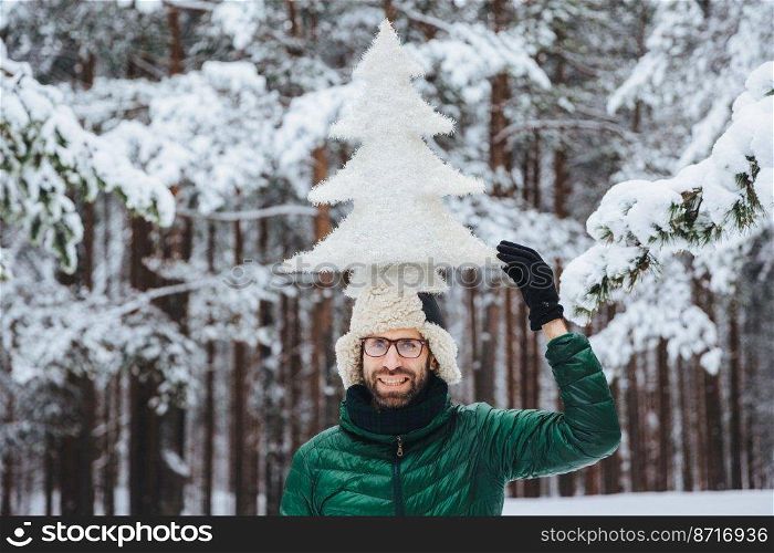Funny bearded male with beard, dressed in warm winter clothes, holds artificial fir tree on head, poses against trees covered with snow, being in good mood. Optimistic man spends time outdoor