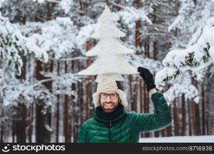 Funny bearded male with beard, dressed in warm winter clothes, holds artificial fir tree on head, poses against trees covered with snow, being in good mood. Optimistic man spends time outdoor