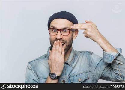 Funny bearded man foolishes indoor, pretends to kiss himself with gun or making suicide gesture, covers mouth with hand, looks joyfully into camera, isolated over grey background. Male foolishes