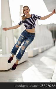 Funny blonde woman jumping in urban background. Beautiful young girl wearing striped t-shirt and blue jeans in the street. Pretty russian female with pigtail.