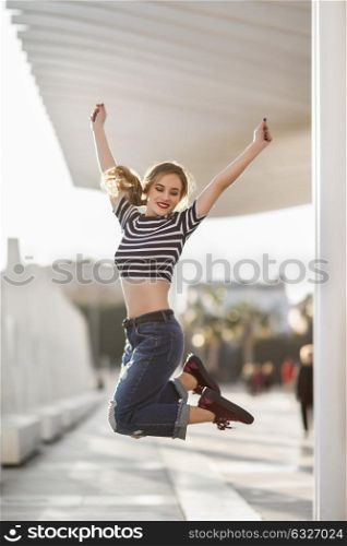 Funny blonde woman jumping in urban background. Beautiful young girl wearing striped t-shirt and blue jeans in the street. Pretty russian female with pigtail.