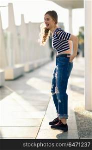 Funny blonde woman laughing in urban background. . Funny blonde woman laughing in urban background. Beautiful young girl wearing striped t-shirt and blue jeans in the street. Pretty russian female with pigtail.