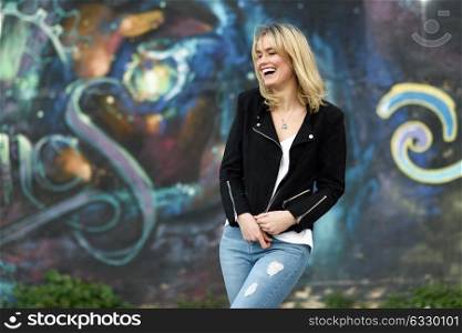 Funny blonde woman laughing in urban background. Young girl wearing black zipper jacket and blue jeans trousers standing in the street. Pretty female with straight hair hairstyle and blue eyes.