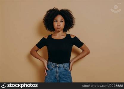 Funny carefree young dark skinned female in casual outfit blowing puffing out her cheeks and having fun indoors against nude studio wall background. People and fun concept, face expressions. Funny carefree young dark skinned female puffing out her cheeks against nude studio wall background