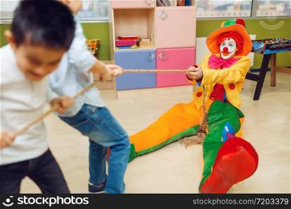 Funny clown and joyful little boys playing tug of war together. Birthday party celebrating in playroom, baby holiday in playground. Childhood happiness, childish leisure. Clown and joyful little boys playing tug of war