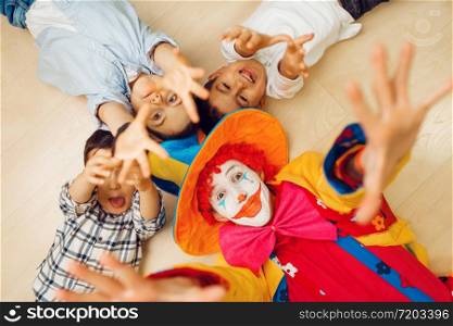 Funny clown and little children lying on the floor and pull their hands into the camera, top view. Birthday celebrating, baby holiday in playground. Childhood happiness, childish leisure. Clown and children pull hands into the camera