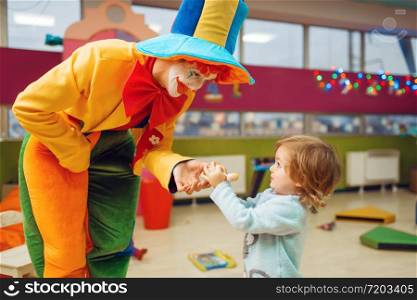 Funny clown gives out lollipop to happy little girl, friendship forever. Birthday party celebrating in playroom, baby holiday in playground. Childhood happiness, childish leisure. Funny clown gives out lollipop to little girl