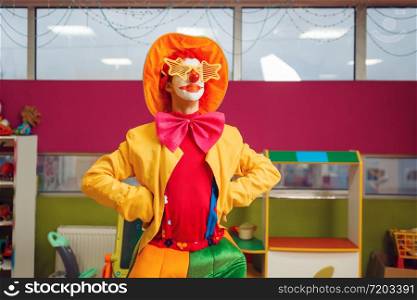 Funny clown in star glasses poses in children&rsquo;s area. Birthday party celebrating in playroom, baby holiday in playground. Childhood happiness, childish leisure, entertainment with animator. Funny clown in star glasses