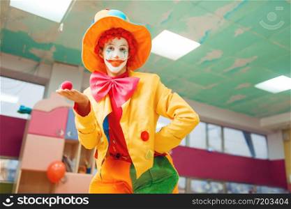 Funny clown poses with red nose in hand. Birthday party celebrating in playroom, baby holiday in playground. Childhood happiness, childish leisure with animator. Funny clown poses with red nose in hand