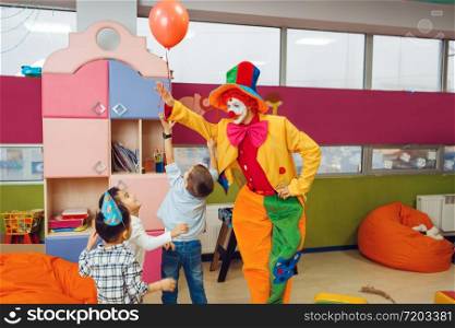Funny clown shows tricks with balloon to surprised children. Birthday party celebrating in playroom, baby holiday in playground. Childhood happiness, childish leisure. Funny clown shows tricks to surprised children