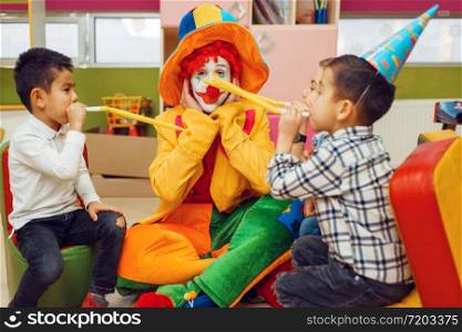 Funny clown with cheerful children blows into the tubes together. Birthday party celebrating in playroom, baby holiday in playground. Childhood happiness, childish leisure. Funny clown with children blows into the tubes