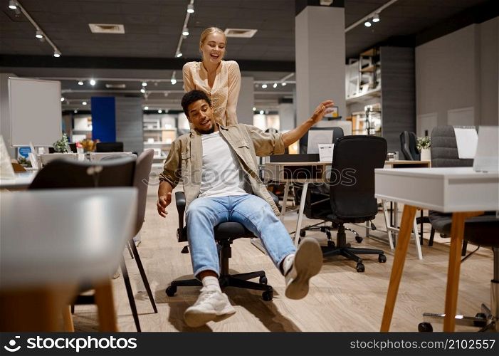 Funny couple riding office chair at furniture shop. Man and woman of different skin color having fun. Funny couple riding office chair at shop