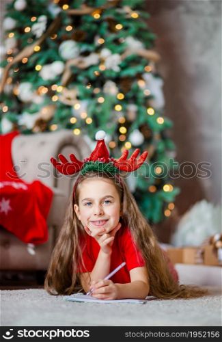 Funny cute caucasian girl left-handed writes a letter to Santa Claus near the Christmas tree. Happy childhood, time of miracles and desires. Merry Christmas.. Funny cute caucasian girl left-handed writes a letter to Santa Claus near the Christmas tree. Happy childhood, time of miracles and desires.