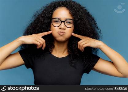 Funny dark skinned woman with curly hair, presses cheeks, holds breath, blows cheeks, wears optical glasses and casual t shirt, isolated over blue background. Female makes grimace at camera.