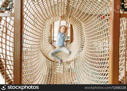 Funny girl plays in rope net, children game center. Excited child having fun on playground indoors. Female kid playing in amusement centre. Funny girl plays in rope net, children game center