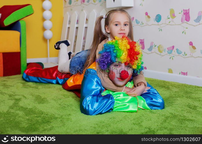 Funny girl sits on a back of the clown with red nose. Colorful sofa, nesting box and fence on the background.
