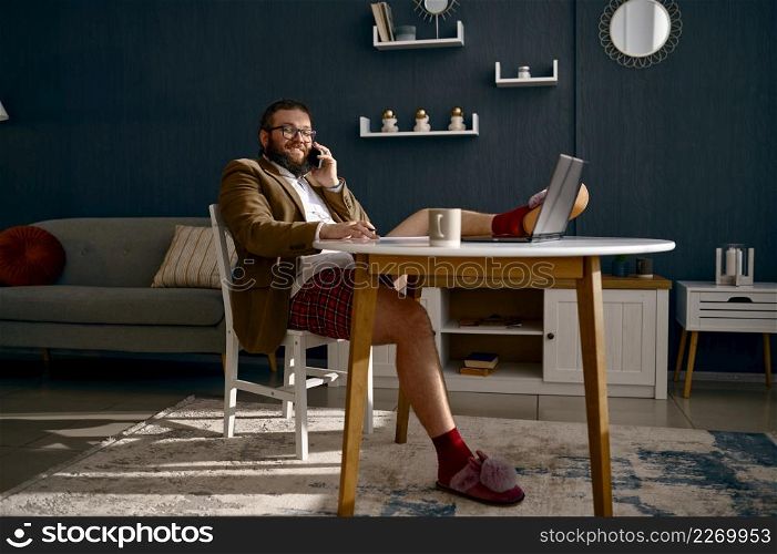 Funny hipster man working at home office. Freelancer in jacket, slipper talking phone sit at desk. Man in jacket and slipper talking phone