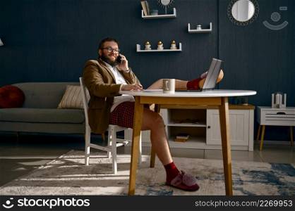 Funny hipster man working at home office. Freelancer in jacket, slipper talking phone sit at desk. Man in jacket and slipper talking phone