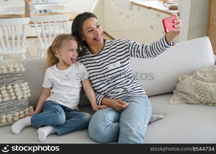 Funny little daughter and young mother showing tongue taking pictures of family, looking at mobile phone camera, happy mom having fun with kid girl, taking selfie together, sitting on couch at home.. Little daughter, mother show tongues taking selfie of family with phone having fun together at home