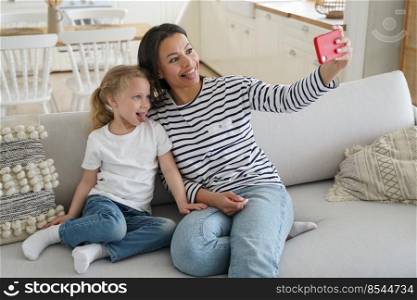 Funny little daughter and young mother showing tongue taking pictures of family, looking at mobile phone camera, happy mom having fun with kid girl, taking selfie together, sitting on couch at home.. Little daughter, mother show tongues taking selfie of family with phone having fun together at home
