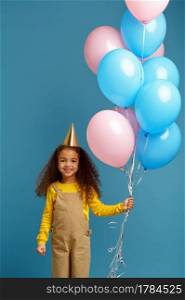 Funny little girl in cap holds a bunch of colorful balloons, blue background. Pretty child got a surprise, event or birthday party celebration. Funny little girl in cap holds a bunch of balloons