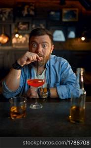 Funny man drinks alcohol coctail at the counter in bar. One male person resting in pub, human emotions, leisure activities, nightlife. Funny man drinks alcohol coctail at counter in bar