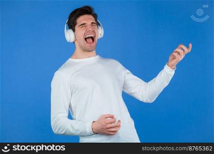 Funny man listening to music with wireless headphones, guy having fun, depicts air guitar playing on blue background. Dance, radio concept. High quality photo. Funny man listening to music with wireless headphones, guy having fun, depicts air guitar playing on blue background. Dance, radio concept.