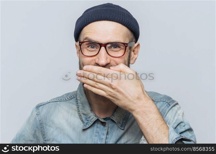 Funny man with attractive look giggles or laughs joyfully, covers mouth with hand, tries to stop feelings, hears anecdote from friend, wears glasses, isolated over grey background. Positive emotions