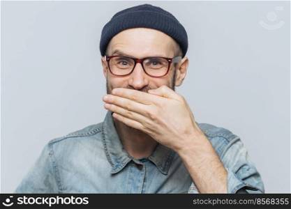 Funny man with attractive look giggles or laughs joyfully, covers mouth with hand, tries to stop feelings, hears anecdote from friend, wears glasses, isolated over grey background. Positive emotions