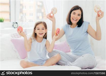 Funny mother and daughter dressed in nightwear, have good mood in morning, hold tasty doughnuts, going to have dessert for breakfast, pose together in bedroom. Family and togetherness concept